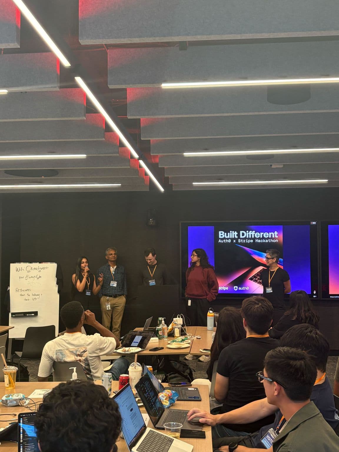 Karishma Mandal speaking into a microphone alongside four other speakers in front of a screen reading Built Different, Auth0 x Stripe Hackathon, with participants at laptops in the foreground.