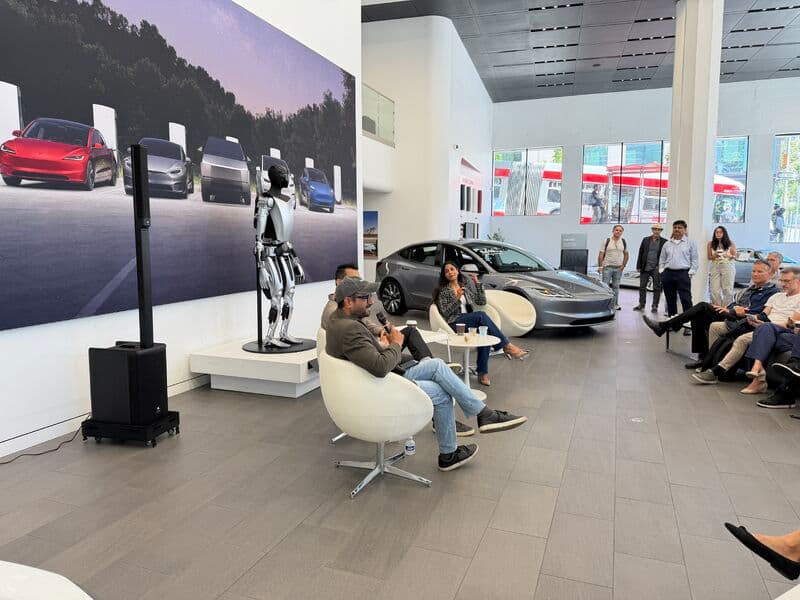 Audience seated and standing around the panel in the Tesla showroom, with a vehicle and the Optimus robot on display.