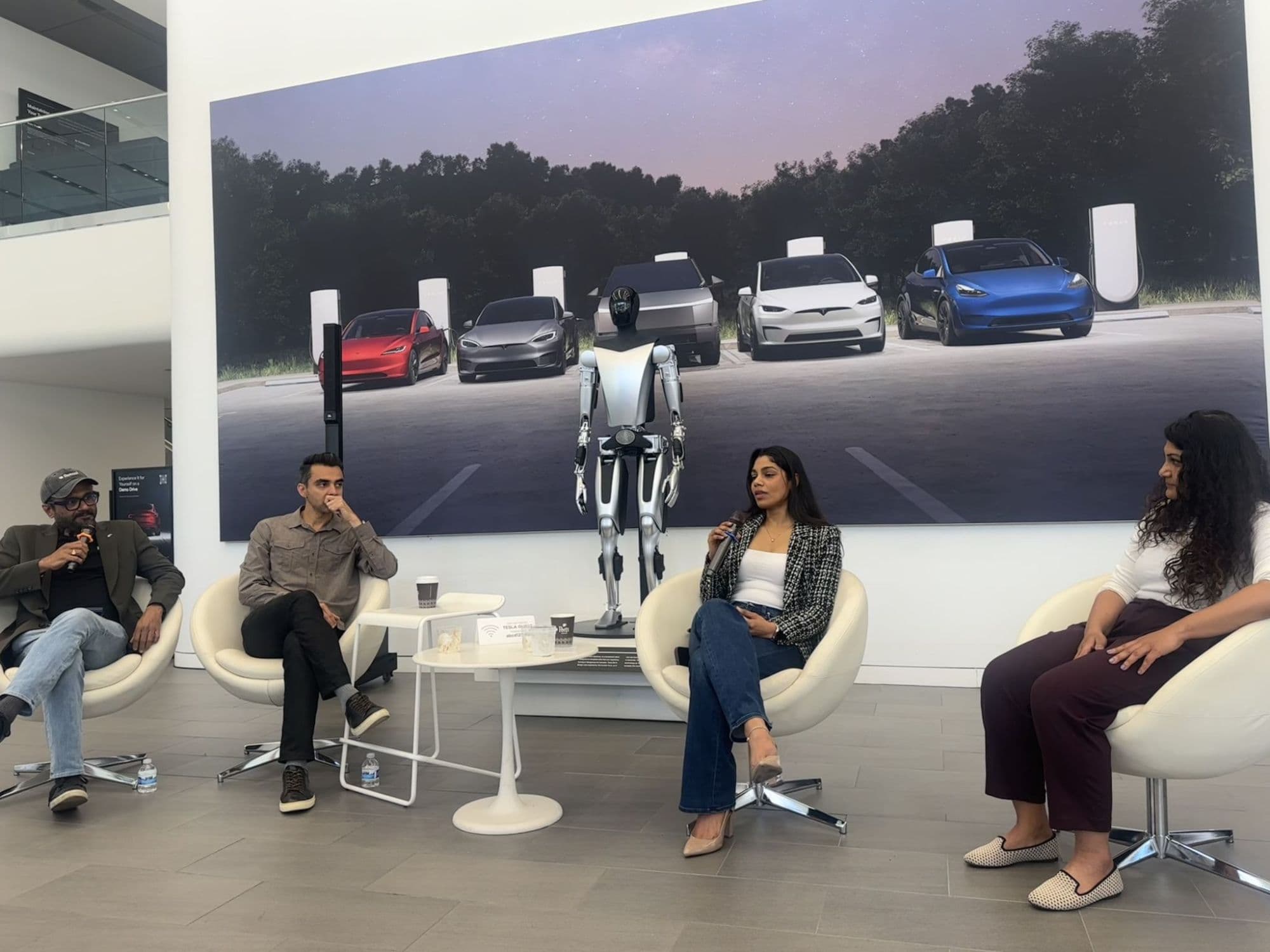 Four panelists seated in white chairs in a Tesla showroom, with an Optimus robot and a wall-sized image of Tesla vehicles behind them; Karishma Mandal is speaking into a microphone.